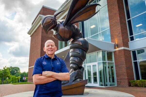 Bill Stewart stands with arms crossed in front of a bronze Hornet statue.