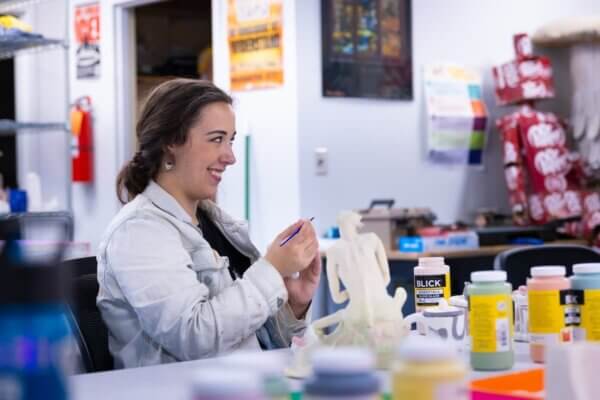 Student in Shenandoah University's Art & Design program smiles and looks away from the camera as works on a small sculpture of a seated human figure.