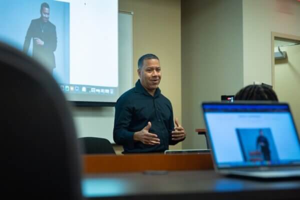 Musician, writer, speaker, entrepreneur and law professor Marcus Johnson talks to Dr. Miles Davis' entrepreneurship ventures class in November 2025. He's standing in front of a screen with his picture on it, which can also be see on a computer's laptop in the foreground. Johnson is wearing all black.