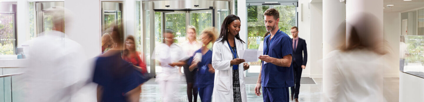 Two healthcare professionals confer together on a busy hospital floor
