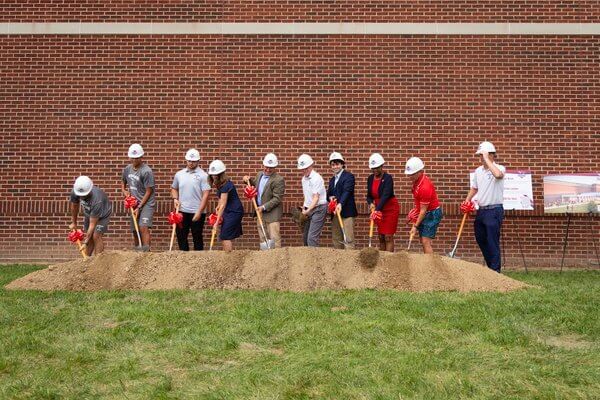 Shenandoah University student-athletes, administrators and coaches wear hardhats and drive shovels into a pile of dirt during a groundbreaking ceremony for an expansion of the Wilkins Athletics and Events Center.