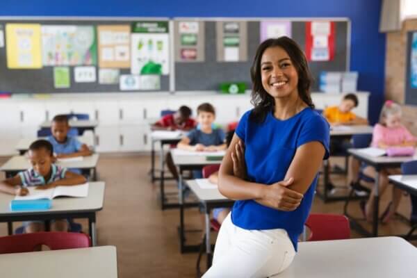 Stock photo of a female-identifying teacher wearing a blue top and white pants leaning against a child-sized desk at the front of an elementary school classroom filled with student studying at their desks.