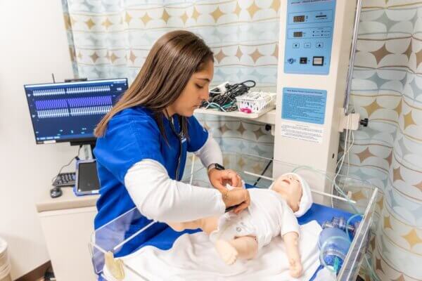 Female-identifying Shenandoah University nursing student wearing blue scrubs trains using an infant manikin in the university's simulation lab.