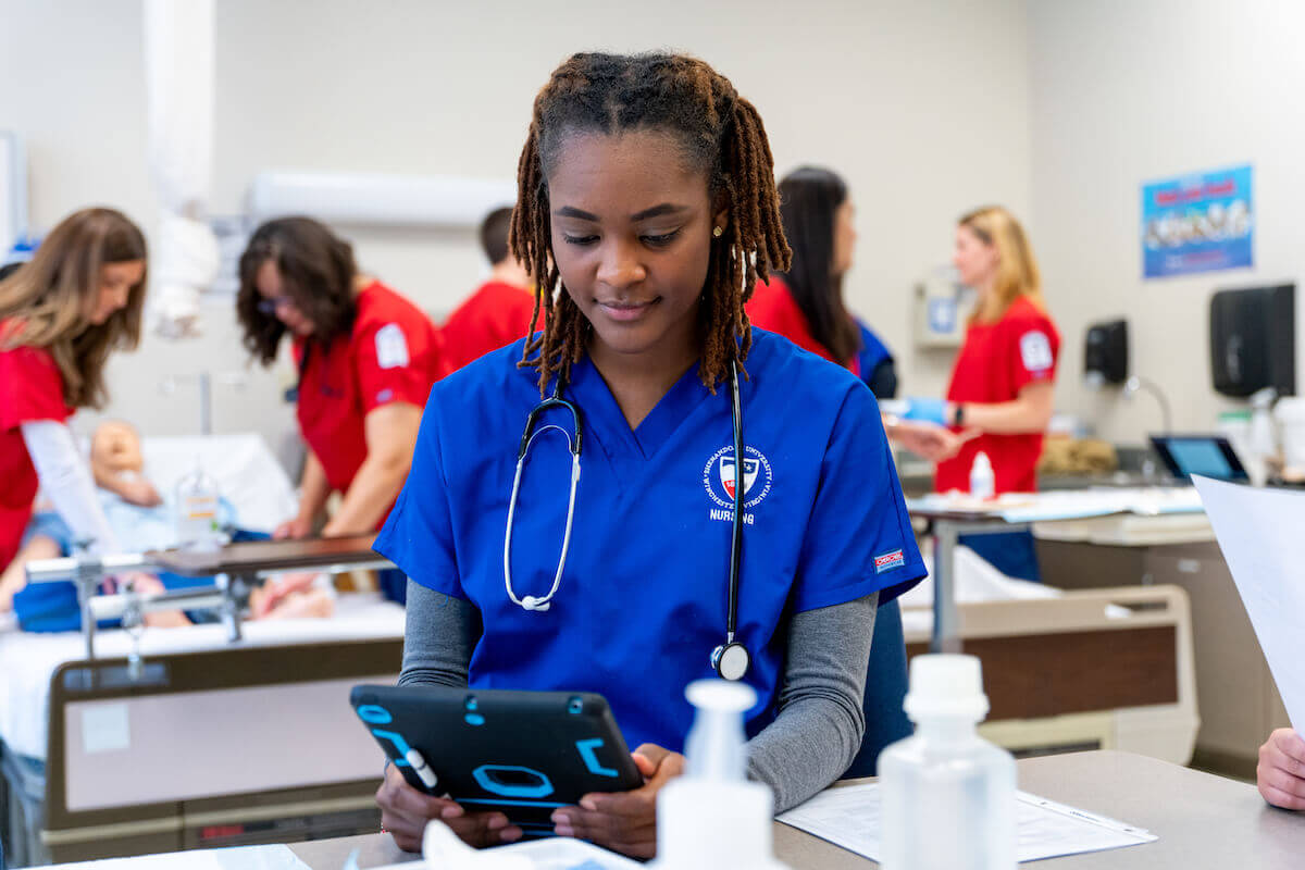 Shenandoah University nursing student in blue scrubs looking at iPad in simulation lab.