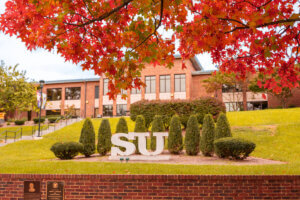 Shenandoah University quad in fall with red maple in the foreground and SU initial statue in center of the photo, in front of Wilkins Administration Building.