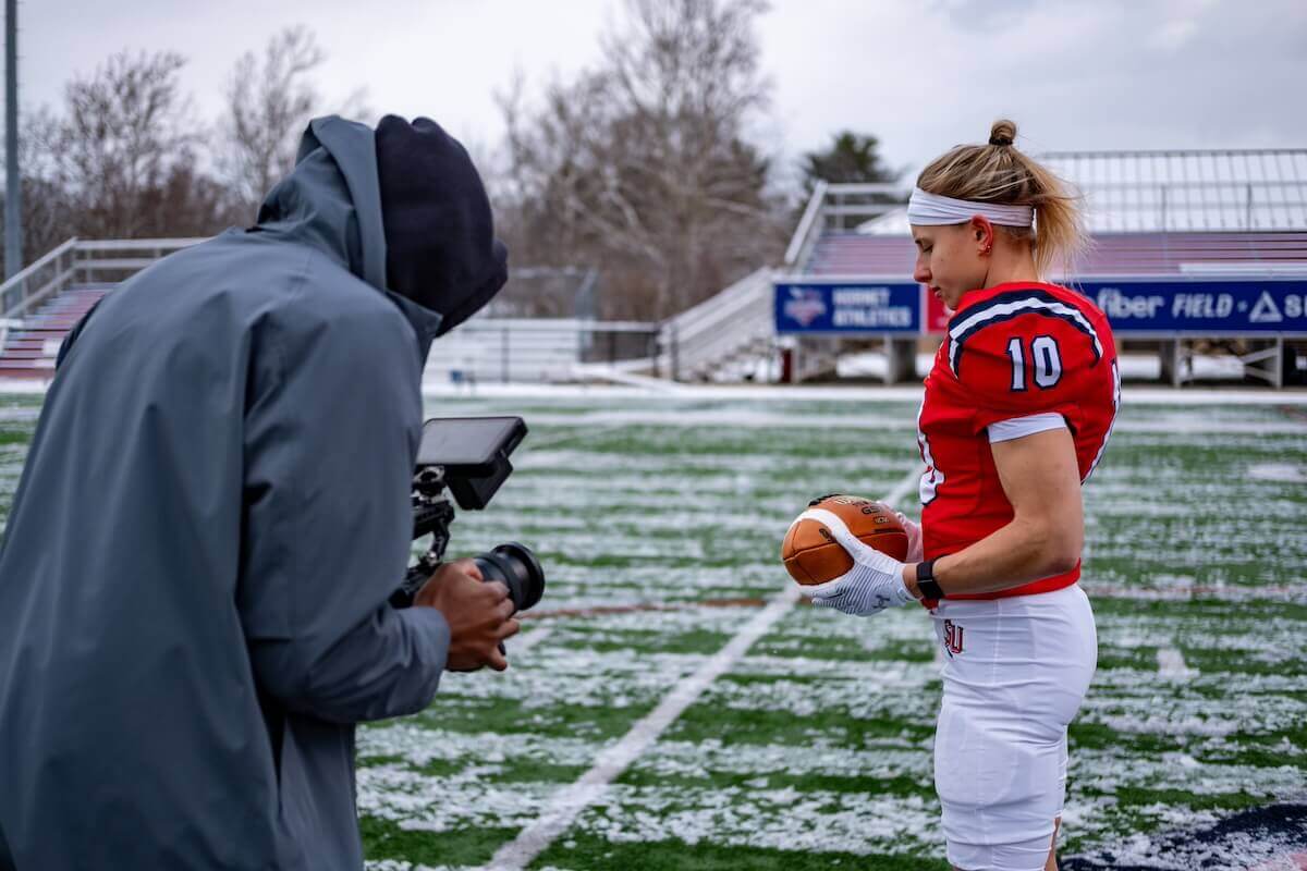 Haley Van Voorhis in a red Shenandoah University football uniform holding a football on a football field while a videographer records footage.