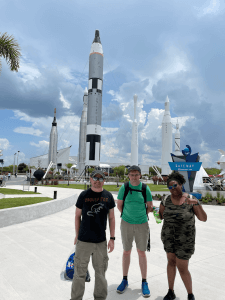 Shenandoah University's Wes Brown, immersive technology specialist and Duasiane’ Benjamin, SCiL operation manager, and VR design student Cole Herndon stand in front of the rocket park at Kennedy Space Center