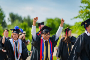 A Shenandoah University graduate raises both arms in celebration as they walk from Shentel Stadium to the Wilkins Athletics and Events Center for Commencement 2023