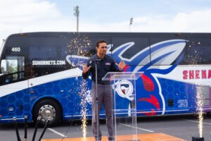 Shenandoah University Athletic Director DeShon Foxx gestures at a podium with the newly designed bus in the background.