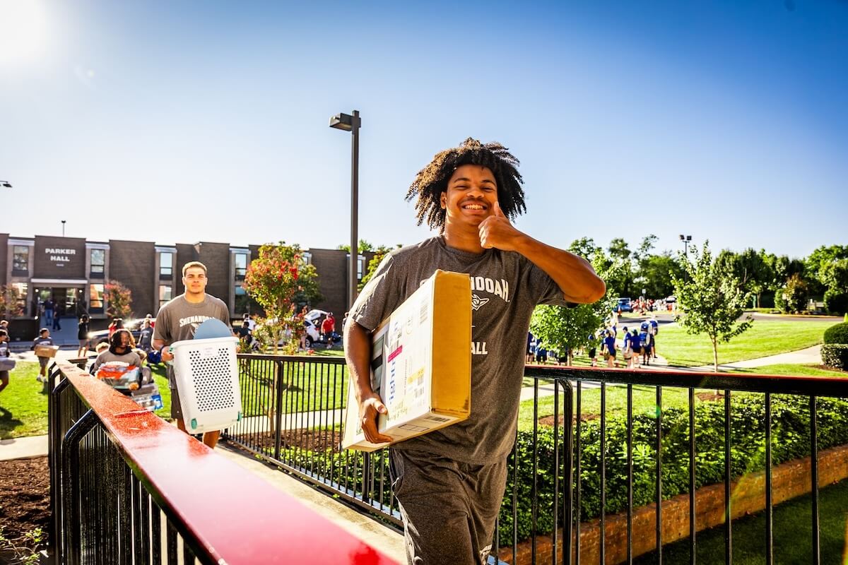 A Shenandoah University student-athlete carries items into a residence hall while giving a thumbs up.
