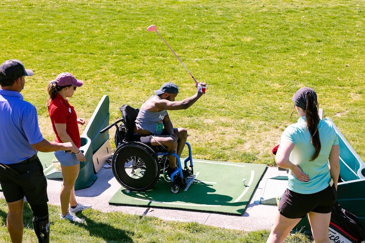 A man in a wheelchair swings a golf club in a Shenandoah University MoveU! adaptive golf clinic, while surrounded by people ready to assist.
