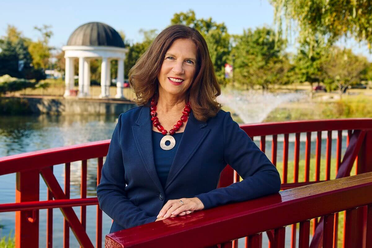 Shenandoah University Tracy Fitzsimmons, Ph.D. wearing a blue outfit and leaning agains the railing of a red bridge on Shenandoah's Winchester main campus, with the Sarah's Glen gazebo visible in the background.