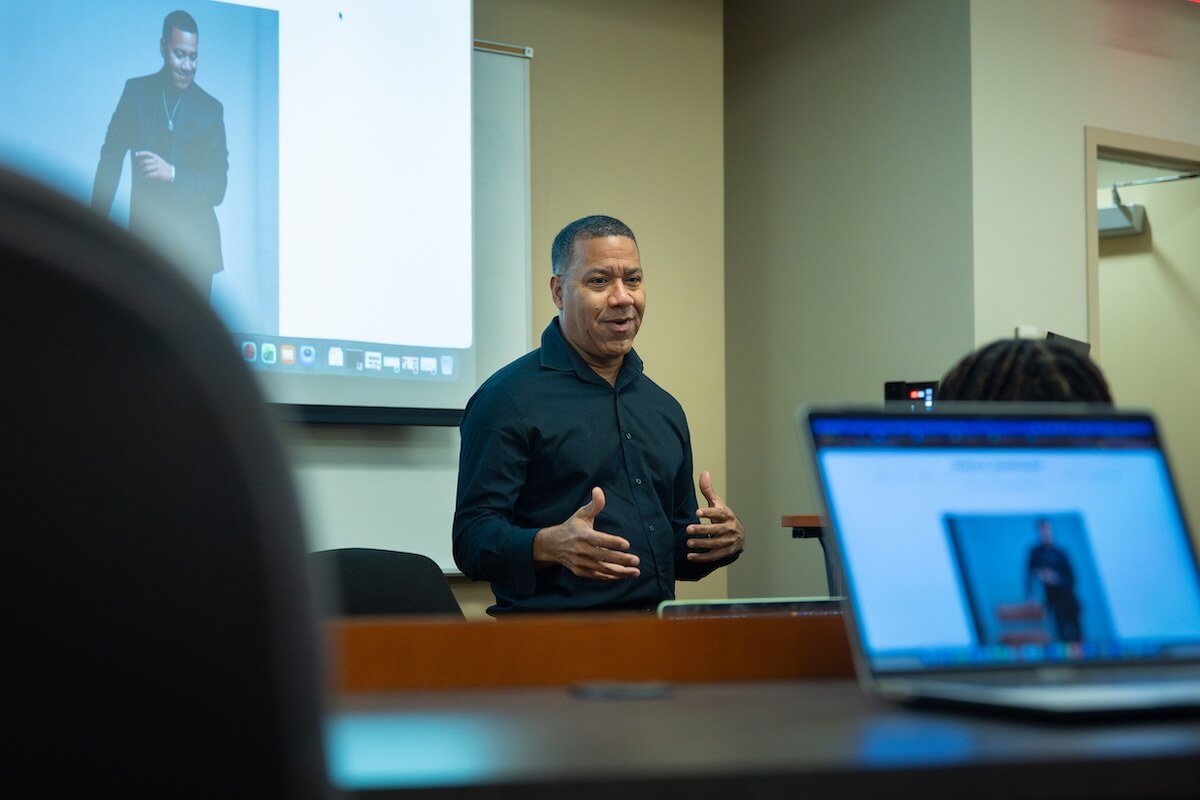Musician, writer, speaker, entrepreneur and law professor Marcus Johnson talks to Dr. Miles Davis' entrepreneurship ventures class in November 2025. He's standing in front of a screen with his picture on it, which can also be see on a computer's laptop in the foreground. Johnson is wearing all black.