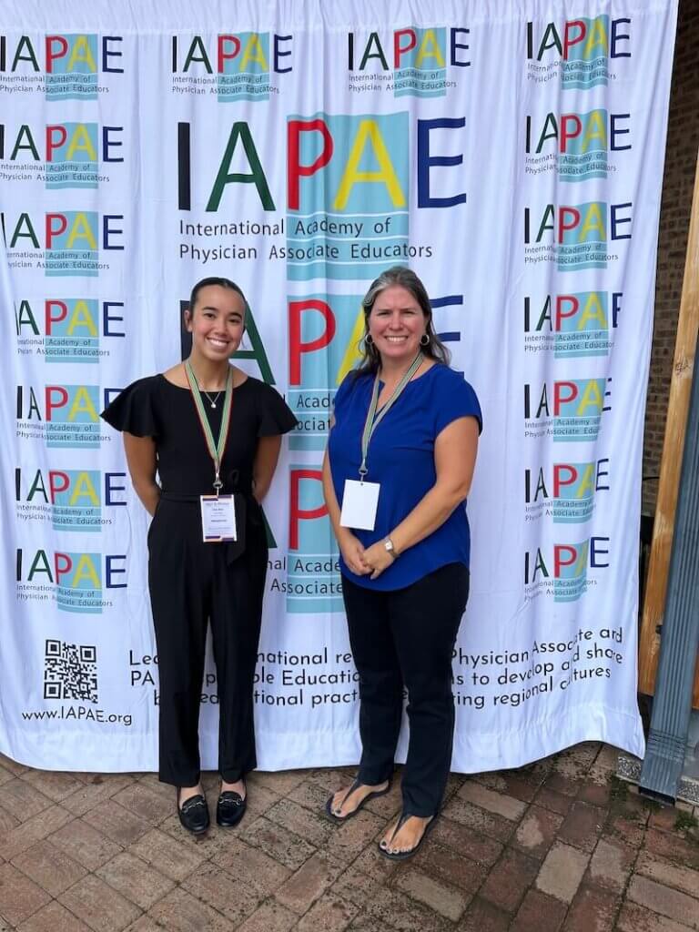 Zoe Star and Camilla Hollen stand in front of a banner for the IAPAE Conference.