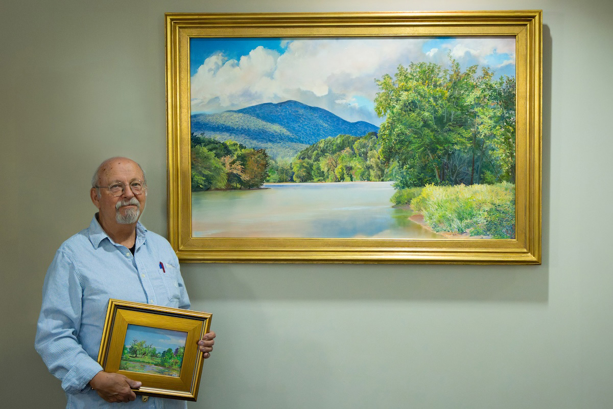 Artist Andrei Kushnir poses with a large framed oil painting of the Shenandoah River while holding another small painting of the Shenandoah Valley landscape.
