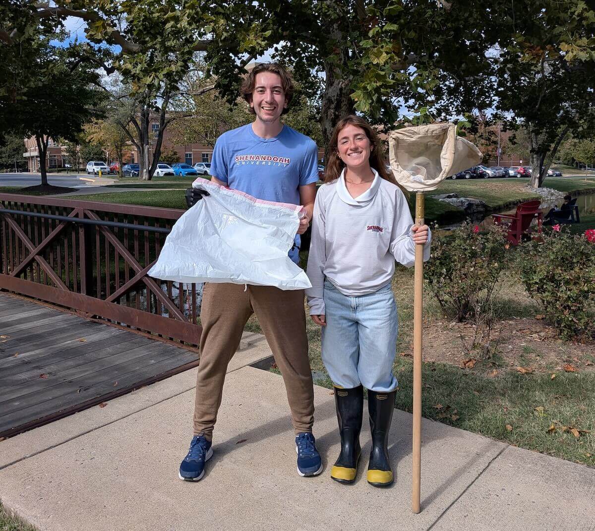 One student holds a trash bag and another is holding a net and wearing rubber boots alongside Abrams Creek.