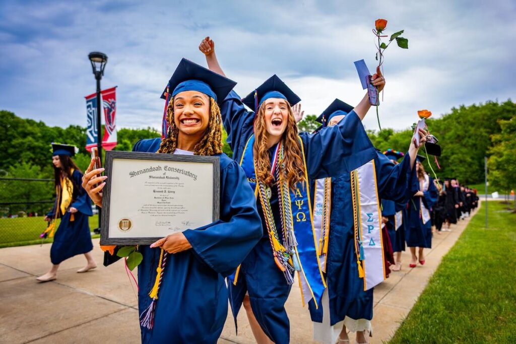 A graduate processes to the commencement ceremony holding a framed copy of their diploma.