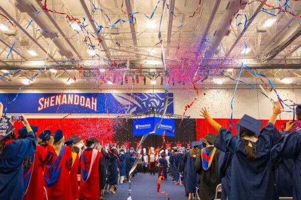 Multicolored streamers fall from the ceiling of the Wilkins Athletics & Events Center at a University Commencement. Graduates in blue caps and gowns and red doctoral robes celebrate.