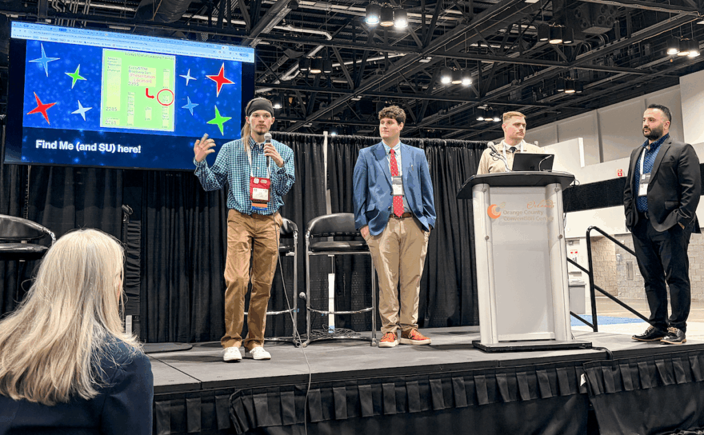 Two students speak on a stage with a display screen behind them.