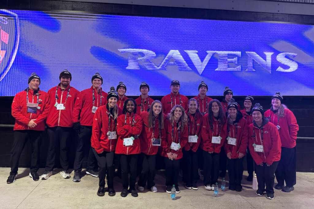 A group of Shenandoah students pose in front of a screen displaying the Baltimore Ravens logo.