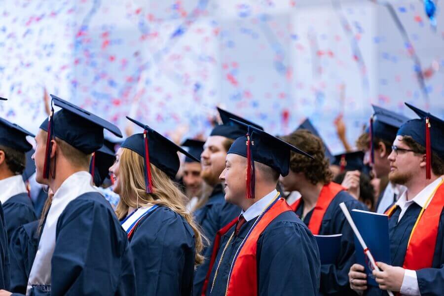 Graduates in caps and gowns stand at commencement with red and blue confetti falling in the background.
