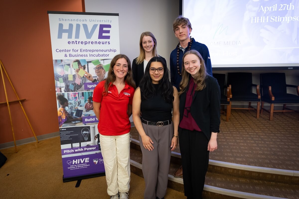 Inaugural HIVE Shenandoah University Pitch Competition participants Abigail Keene ’26, Raquel Anongos ’26, Arizona Fischvogt ’28, Parker Brown ’29, Natalie Pronk ’29 stand in Halpin-Harrison Hall, Stimpson Auditorium, in front of a HIVE pull-up poster focused on entrepreneurship.