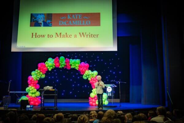 Author Kate DiCamillo in front of a pink and green balloon arch and a slide that says "How to Make a Writer" on stage at Shenandoah University's Ohrstrom-Bryant Theatre in 2026 for the university's Children's Literature Conference's Rally for Reading.