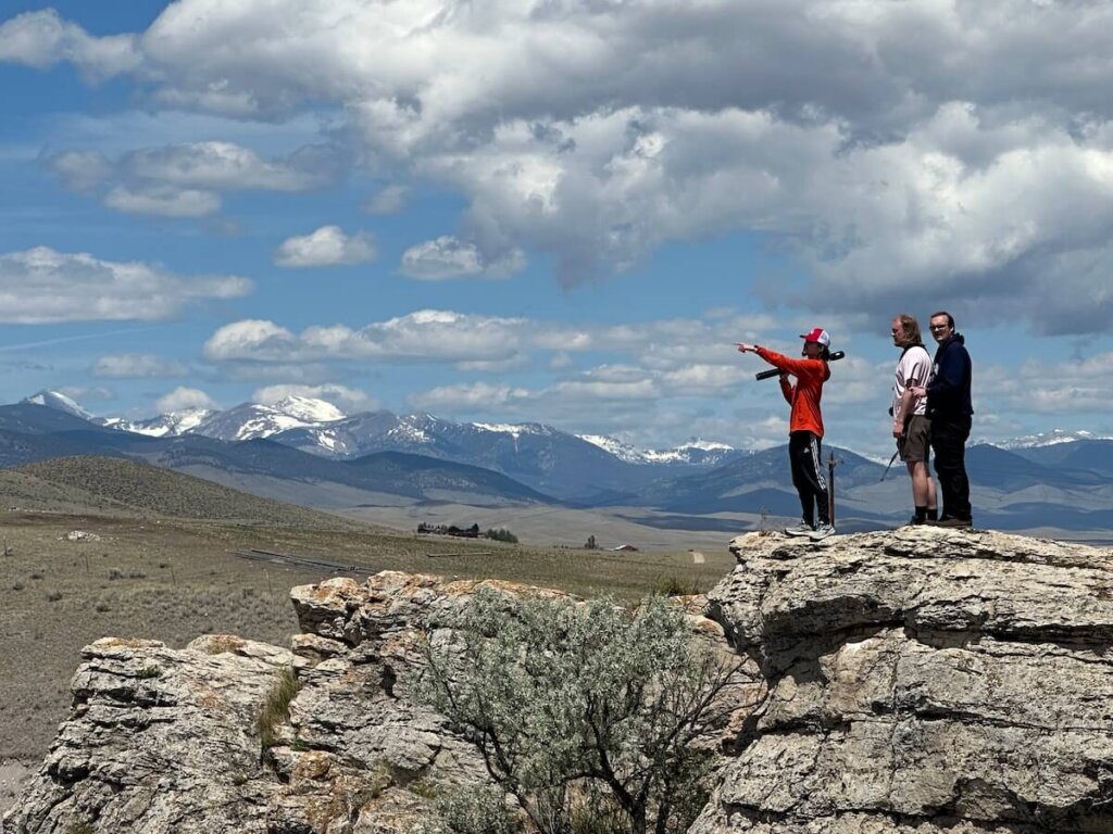 Three students stand on a rocky outcropping with snow-capped mountains in the distance.