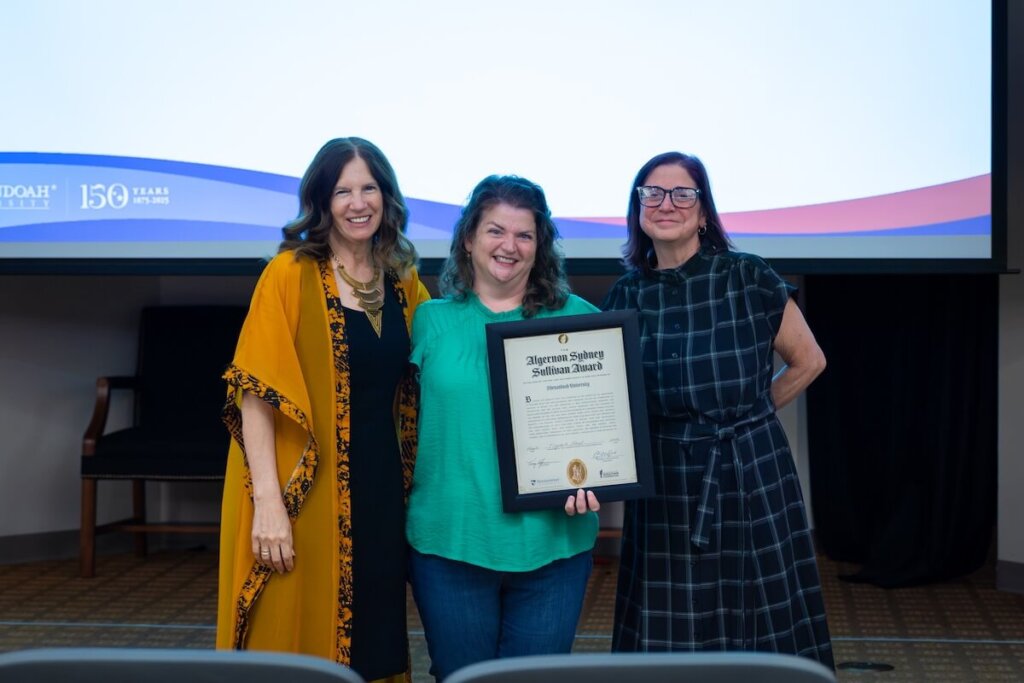 Liz Albert holding a framed copy of her Sullivan Award while standing between Shenandoah University's president and the university chaplain.