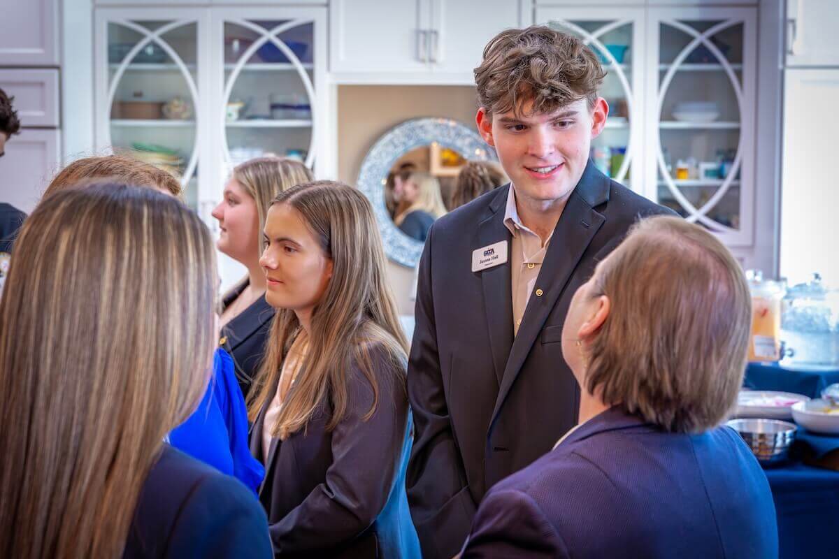 Jaxson Hall '26 (B.S. Psychology-Shenandoah University), wearing a gray suit and name tag, listens to another person at a Shenandoah University Leadership Academy networking event. Other people are present at the event, which looks like it is being held in a home's kitchen/dining area.