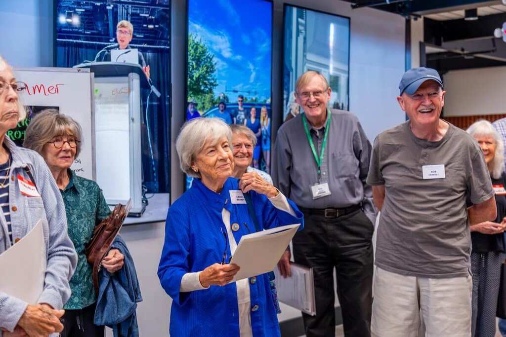 Older adults stand in a hallway as they listen to a speaker who is off-camera.
