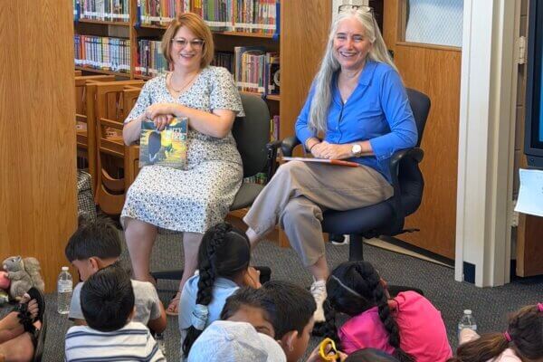 Karen Schaufeld, an author, entrepreneur, philanthropist, lawyer, and ardent early reading advocate, holds her book, "Larry and Bob" as she answers student questions at Sully Elementary School in Loudoun County, Virginia as part of Shenandoah University's Camp Read A Lot. She is smiling, has long silver hair, and is wearing glasses and a blue button-up shirt.