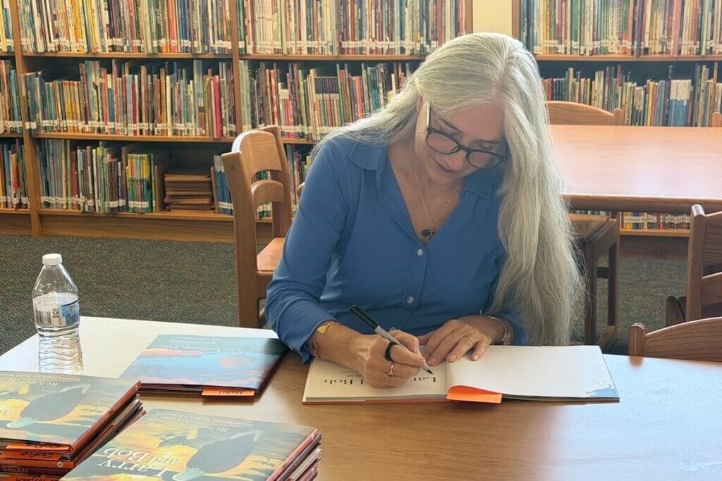 Karen Schaufeld, an author, entrepreneur, philanthropist, lawyer, and ardent early reading advocate, signs her book, "Larry and Bob" at Sully Elementary School in Loudoun County, Virginia as part of Shenandoah University's Camp Read A Lot. She has long silver hair, and is wearing glasses and a blue button-up shirt.