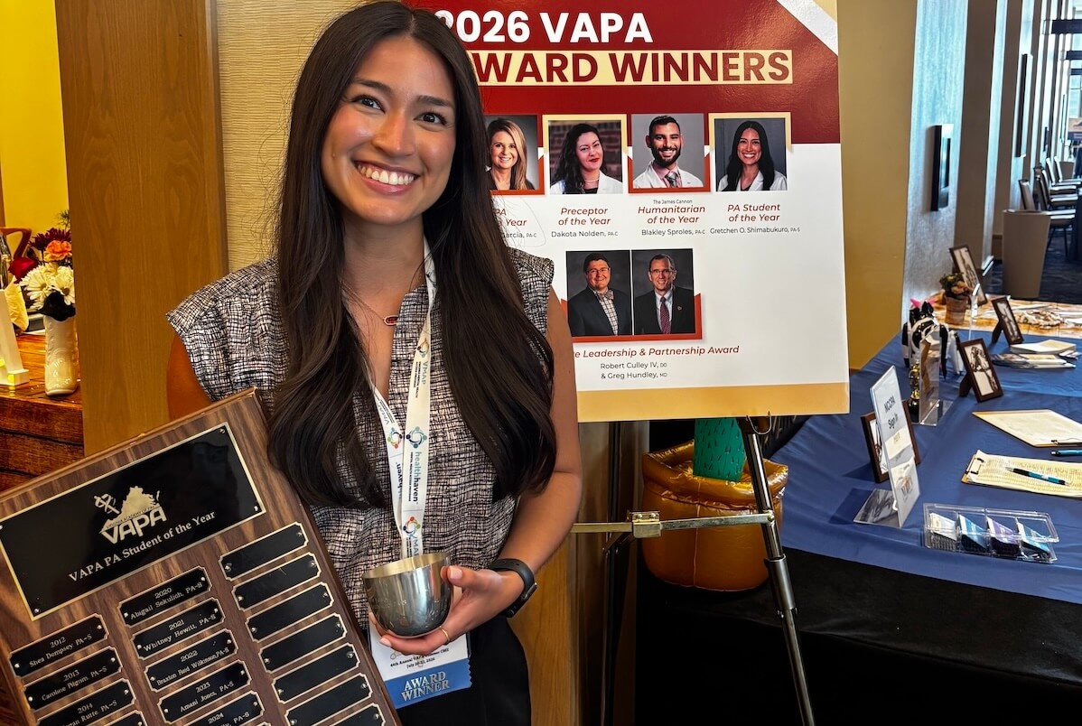Gretchen Olazabal Shimabukuro ’26, a Shenandoah University physician assistant studies student, is the 2026 Virginia Academy of Physician Assistants’ PA Student of the Year, holding the award plaque with her name on it and standing in front of a poster with all the year's VAPA winners on it.