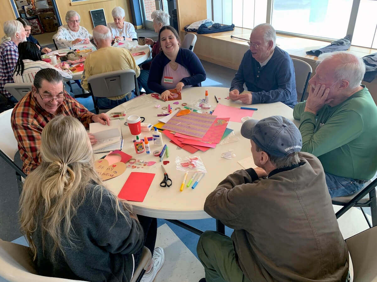 Shenandoah students and older adults sit around a table sharing laughs while crafting.