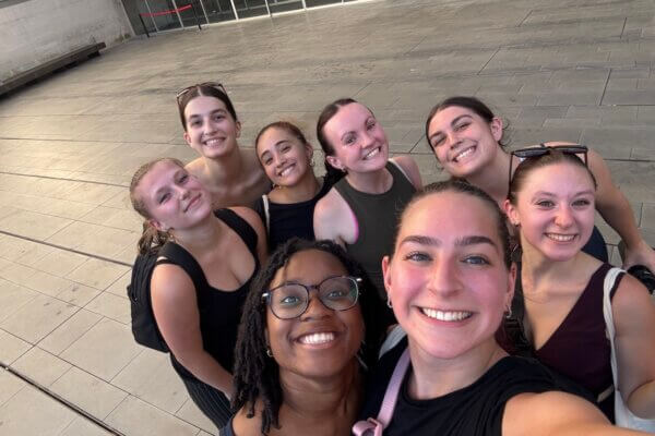 Shenandoah University BFA Dance student Lauren Scott, shown with a pink strap over her right arm, with 7 other dance students in a selfie. All students are wearing black.