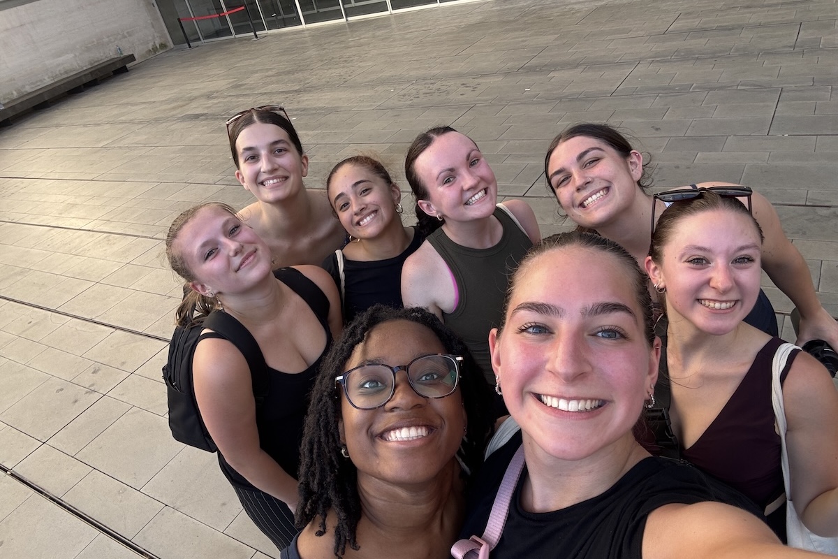 Shenandoah University BFA Dance student Lauren Scott, shown with a pink strap over her right arm, with 7 other dance students in a selfie. All students are wearing black.