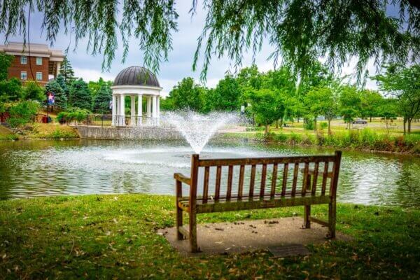 An empy wooden bench beside a pond with a fountain and gazebo in the background.