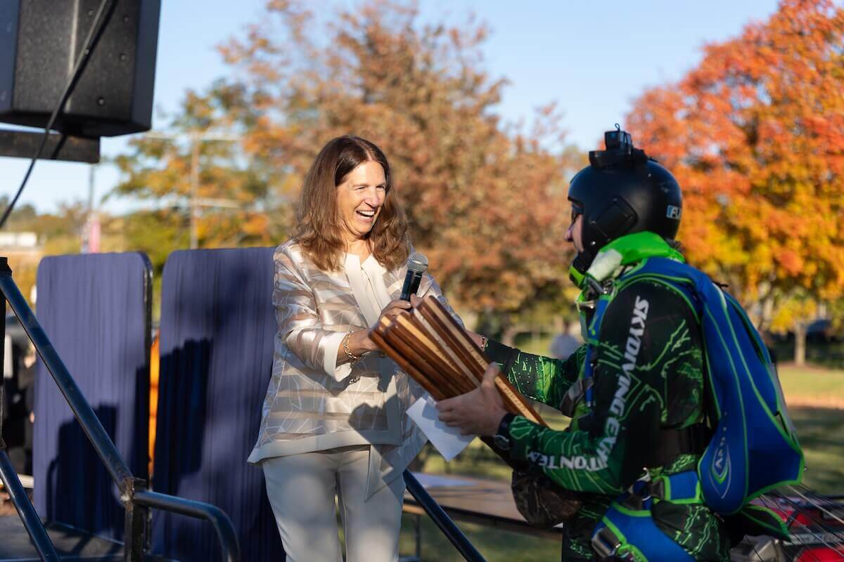 Shenandoah University President Tracy Fitzsimmons receives three custom batons from a skydiver at the podium.