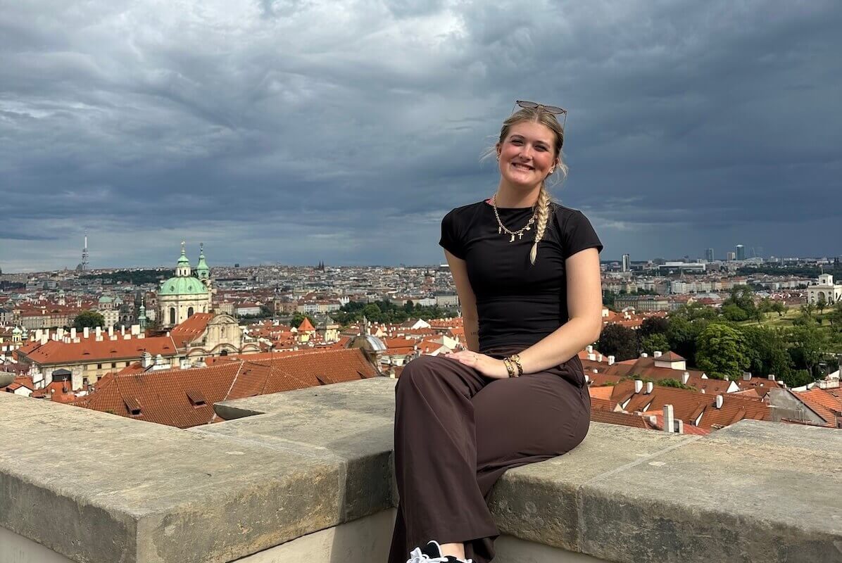 Shenandoah University exercise science major Katerina Deising '27 sitting on a wall in Prague, Czechia, with the city's rooftops behind her, during the summer of 2026, when she interned at the European Box Lacrosse Championships held in the city.