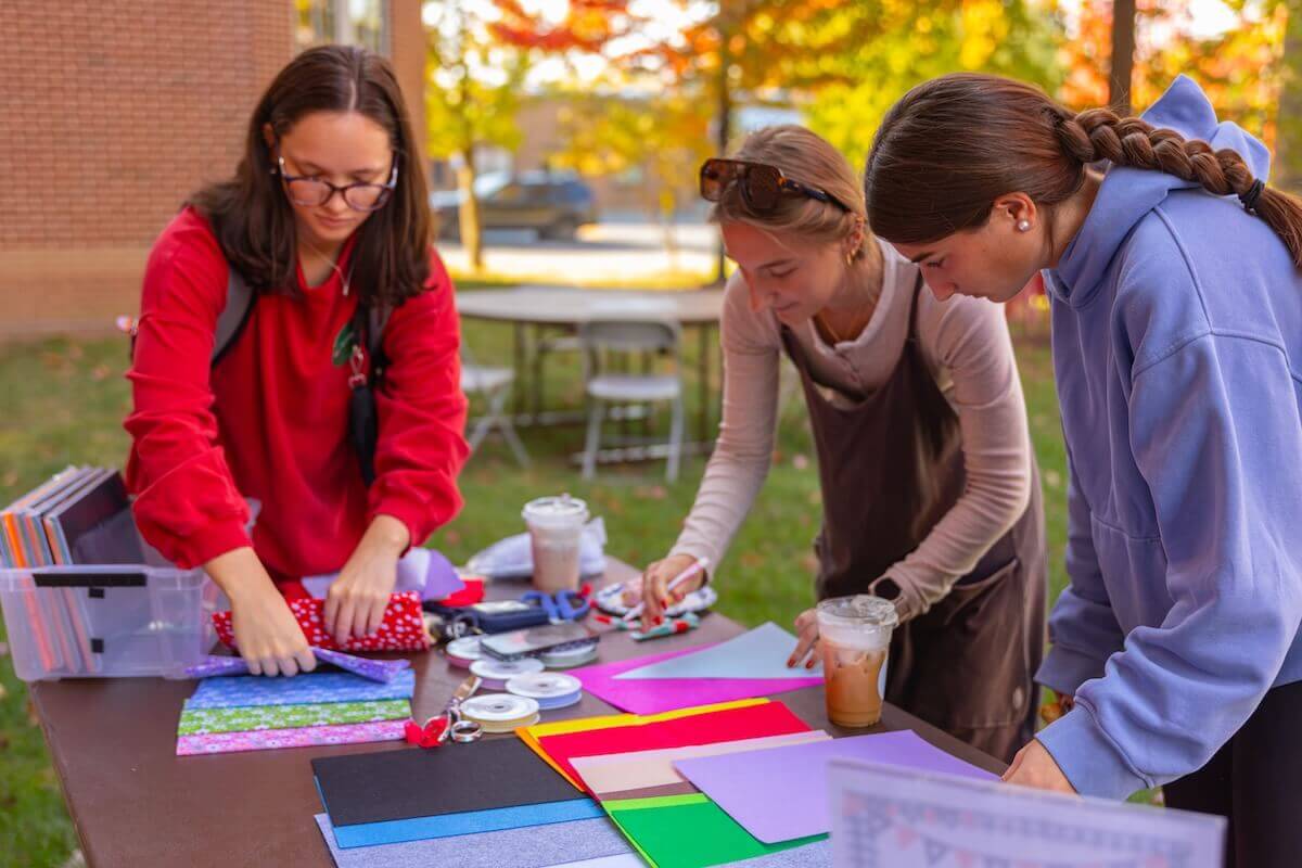 Three students sort through crafting supplies at an outdoor table.