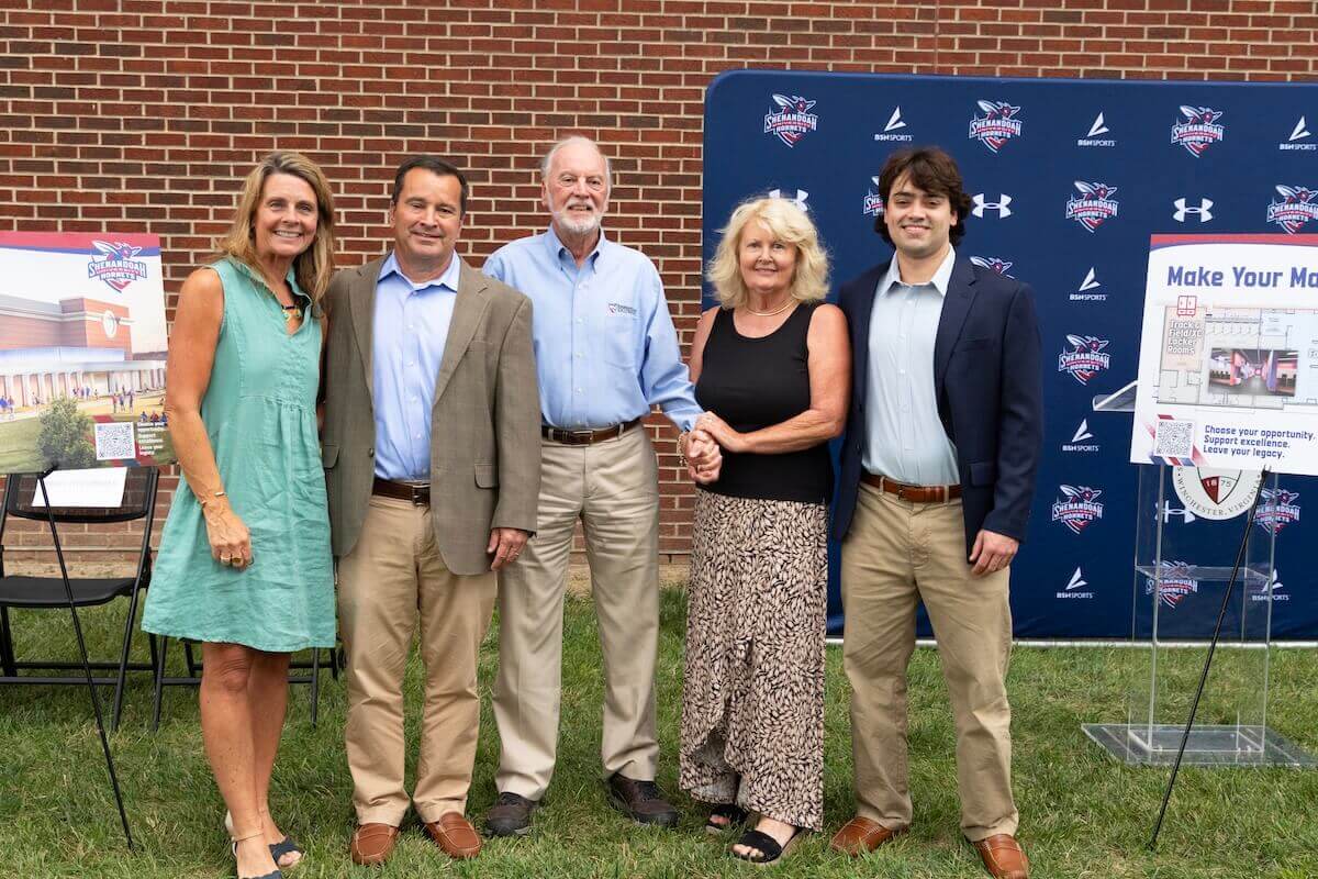 Members of the Wilkins family pose for a group photograph between posters showing renderings of the Wilkins Athletics and Events Center expansion.