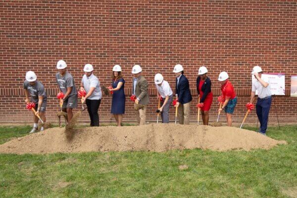 Shenandoah University leadership, coaches and student-athletes wear hardhats and put shovels into a pile of dirt during a ceremonial groundbreaking.