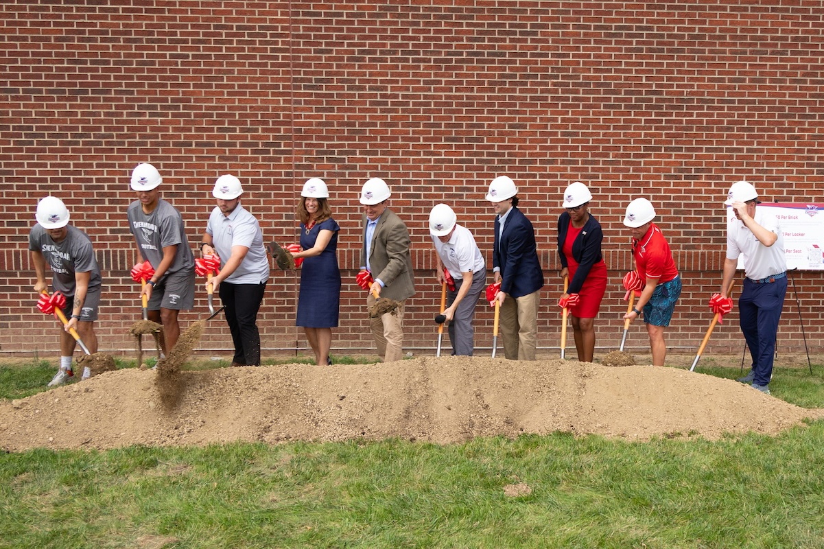Shenandoah University leadership, coaches and student-athletes wear hardhats and put shovels into a pile of dirt during a ceremonial groundbreaking.