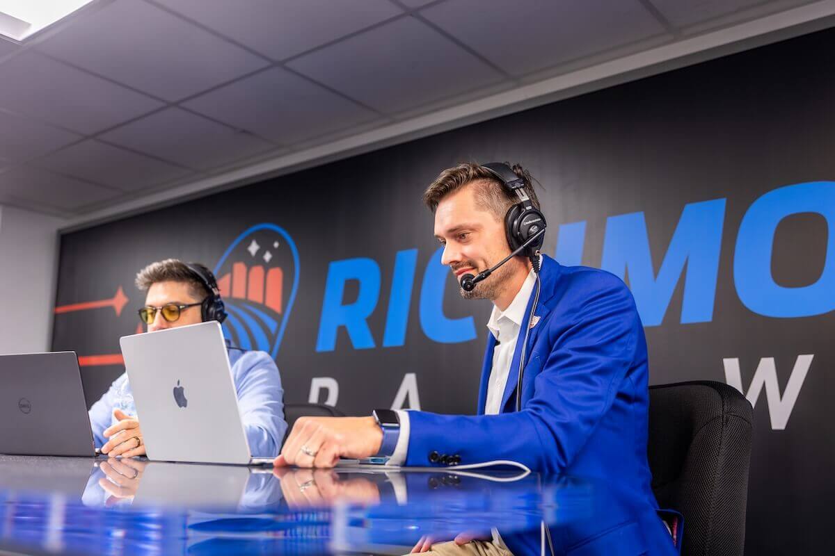 Zander Merle-Smith seated at a desk, wearing a blue suit and a headset while looking at a laptop.