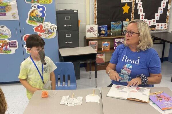 Child wearing fake elephant trunk on his nose and teacher wearing a "Read" T-shirt share an Elephant and Piggie book for parents in an elementary school classroom at a Camp Read A Lot in the summer of 2025.